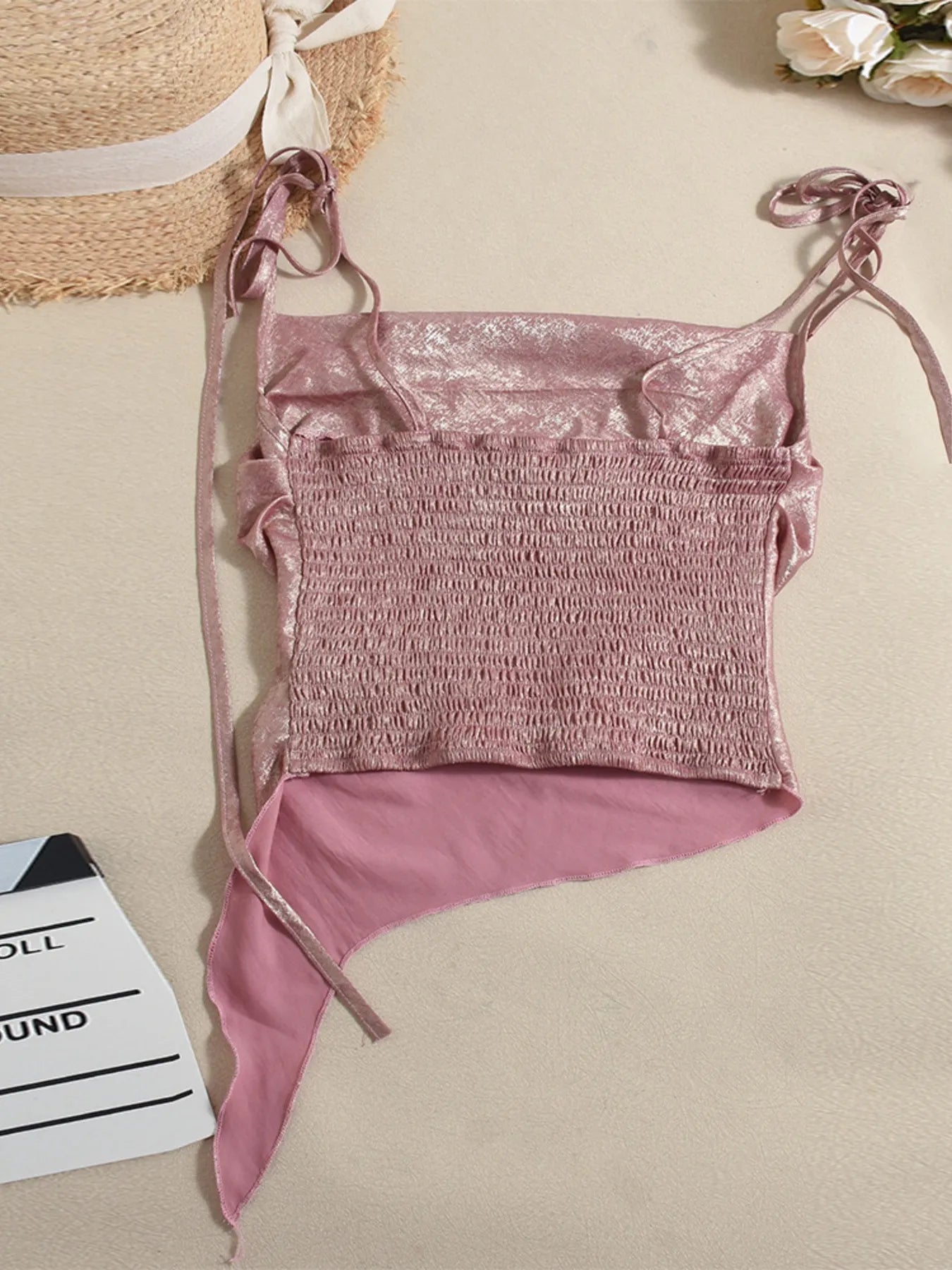 Pink shimmery tie shoulder cami summer top back view on a beige surface with a straw bag and flowers in the background