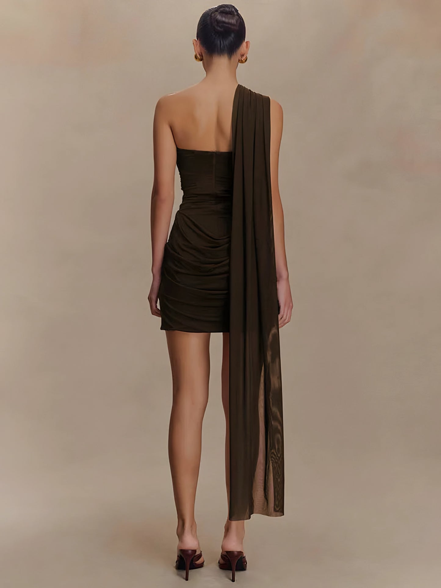 Woman wearing a brown draped one-shoulder mini dress on a beige background back view