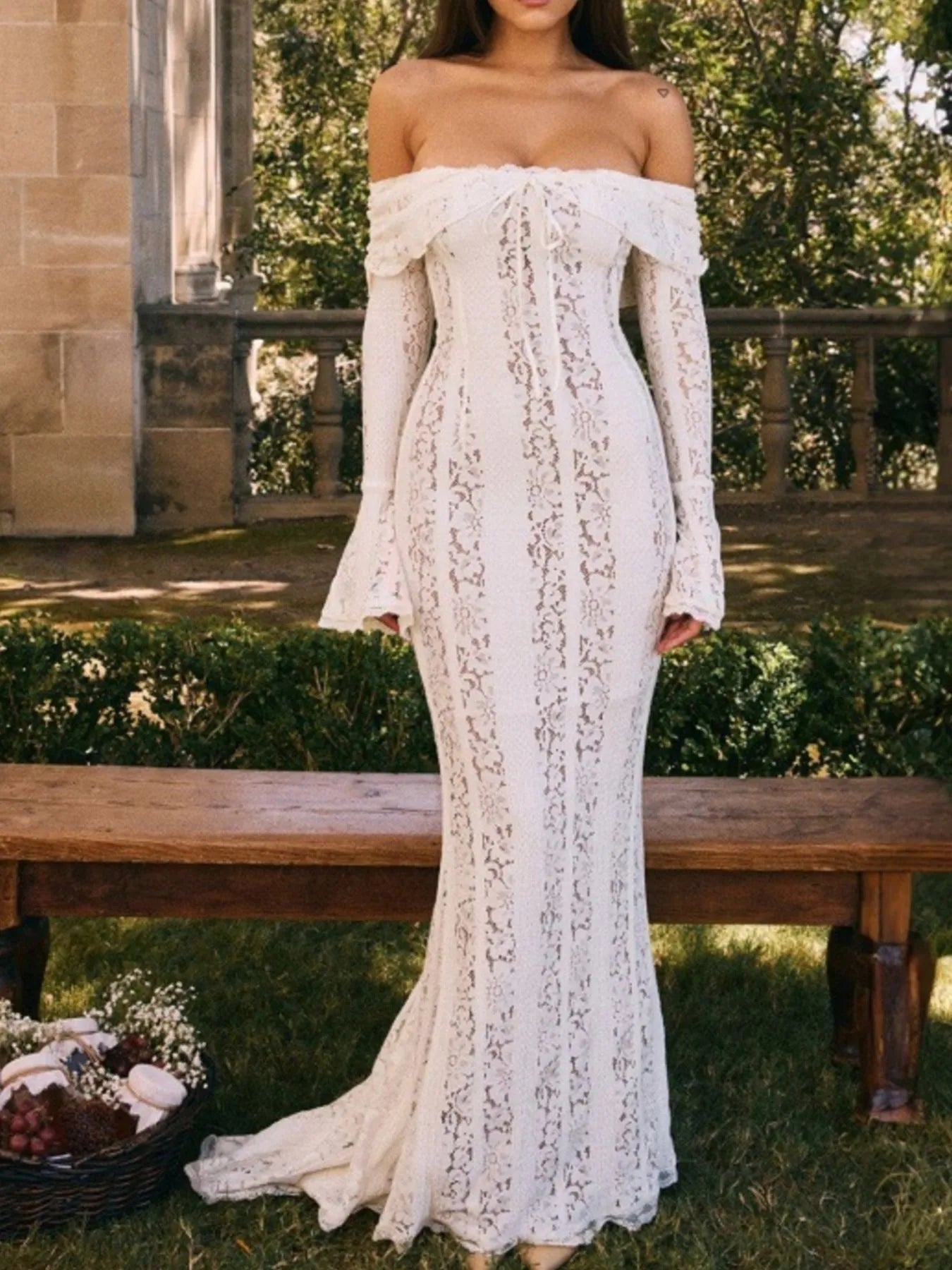 Woman wearing a white lace off-shoulder maxi dress dress standing in front on a wooden bench outdoors.