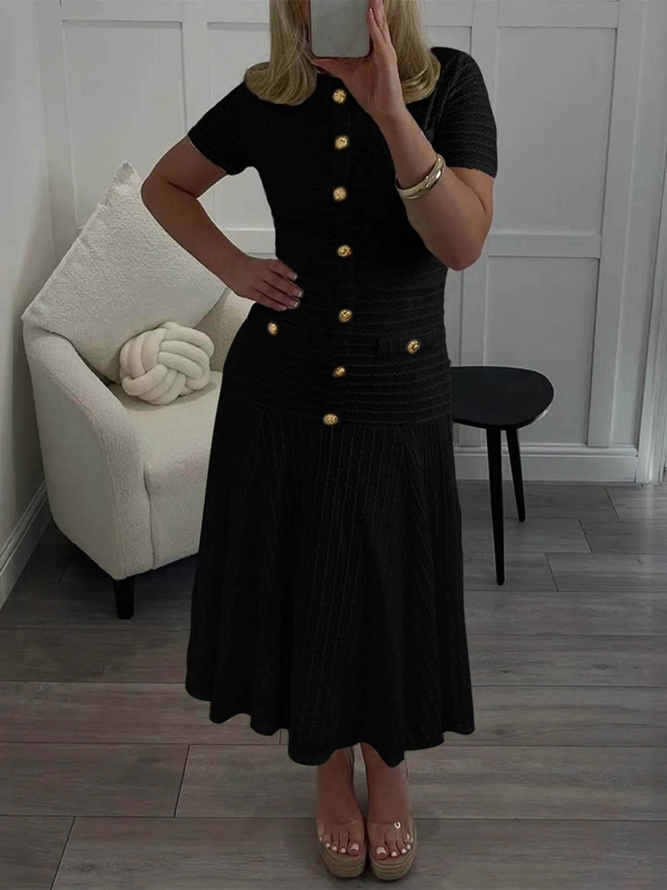 Woman wearing a black casual fitted ribbed midi dress with gold buttons in a room with a white chair and wooden floor.