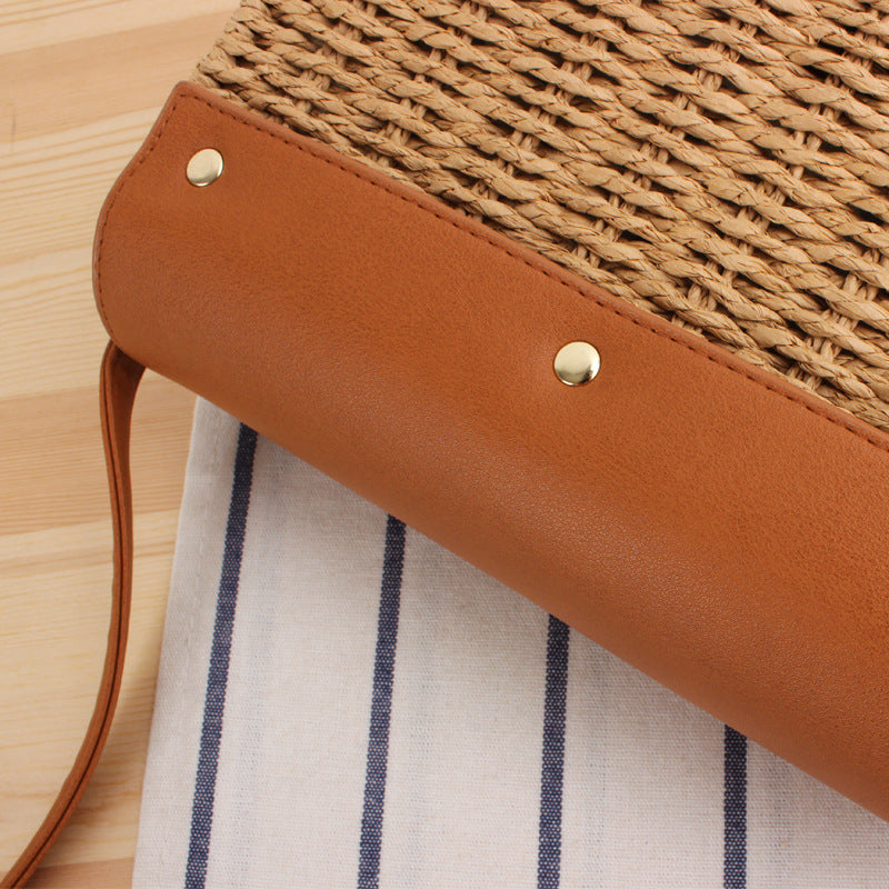 Close-up of a woven straw bag with brown leather strap and flap on a striped fabric background
