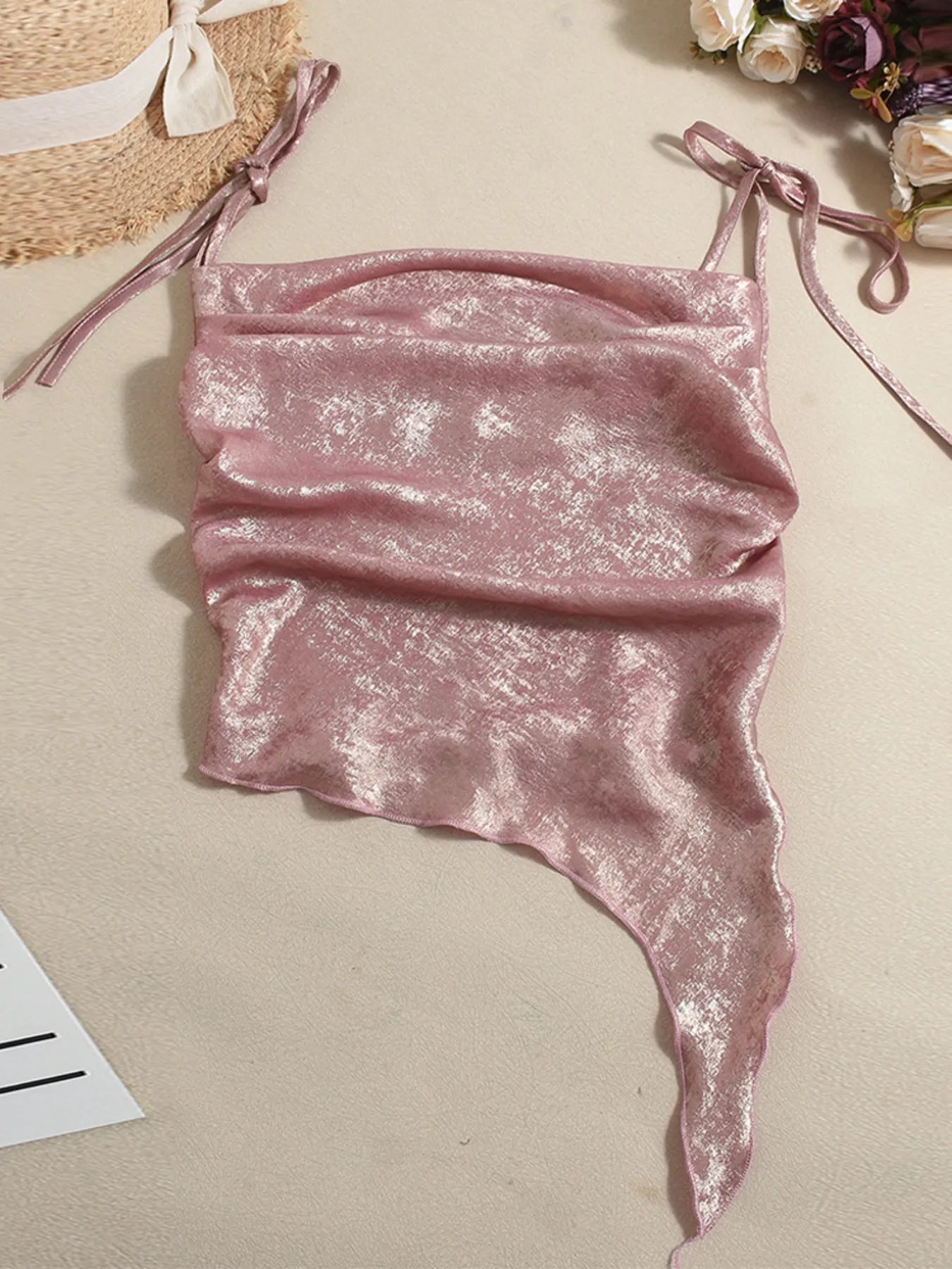 Pink shimmery tie shoulder cami summer top on a beige surface with a straw hat and flowers in the background.