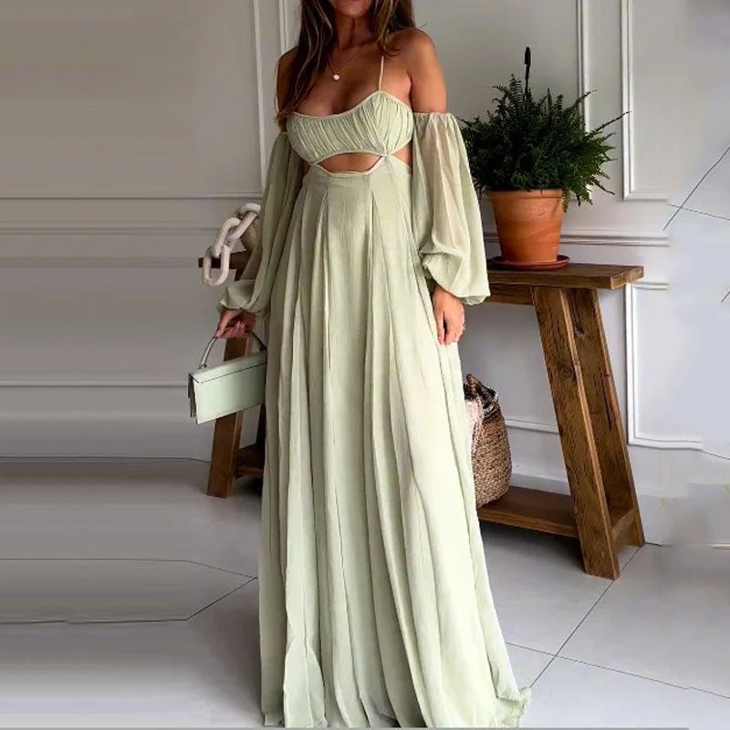 A woman wearing a flowy light green / sage, off-shoulder, backless maxi dress with puff sleeves and a cutout detail standing near a wooden table and a plant.