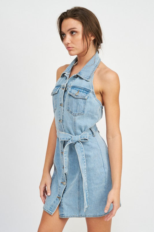 Woman wearing a light blue belted halter denim dress on a white background side view