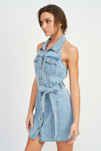 Woman wearing a light blue belted halter denim dress on a white background side view