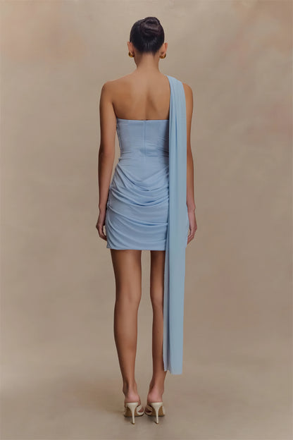 Woman wearing a light blue draped  one-shoulder draped mini dress on a beige background back view