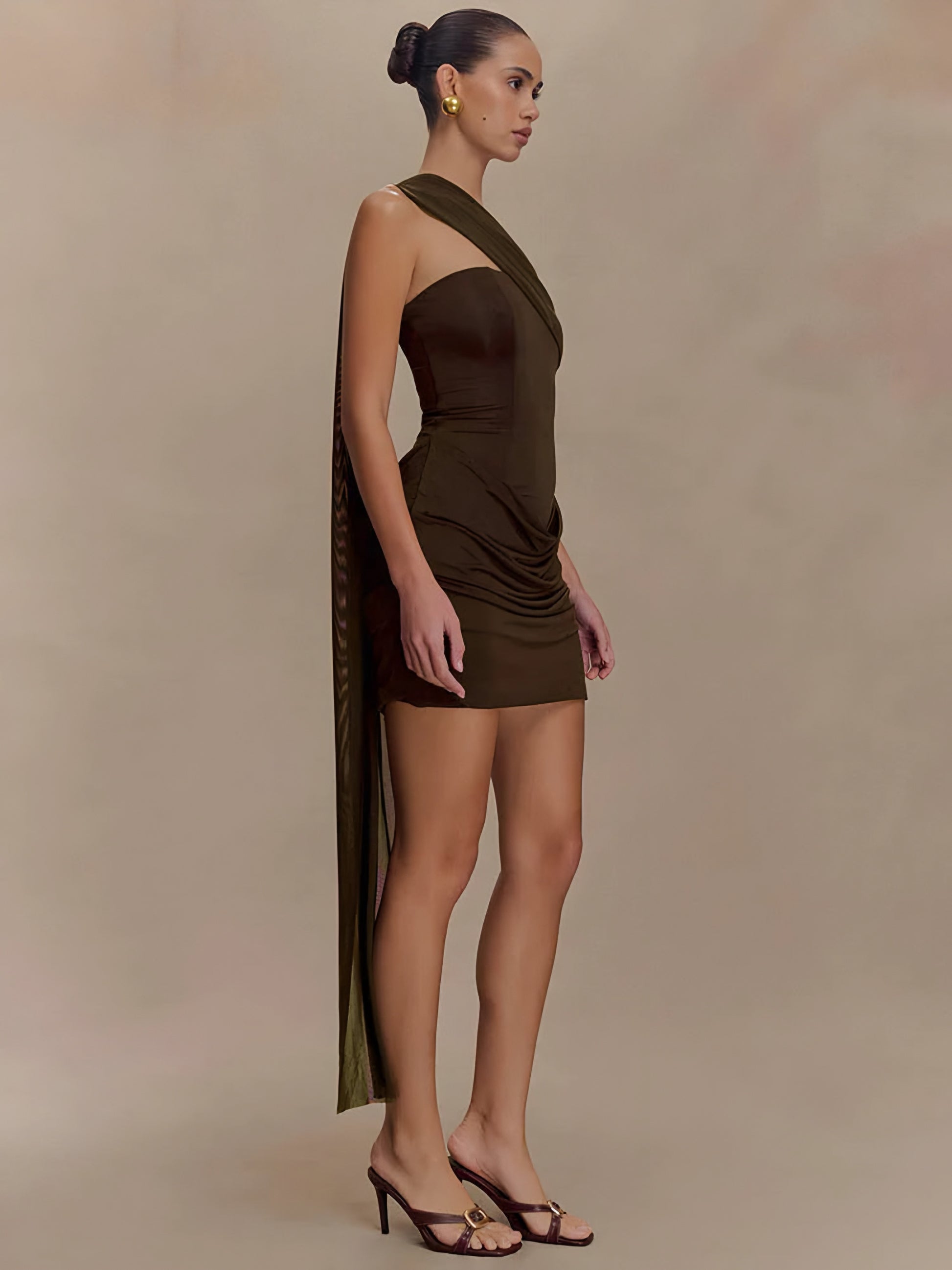 Woman wearing a brown draped one-shoulder mini dress on a beige background side view