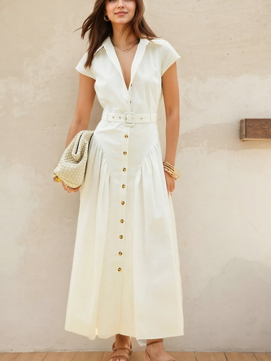 Woman wearing a white button front cap sleeve belted resort chic style casual shirt dress against a beige wall.