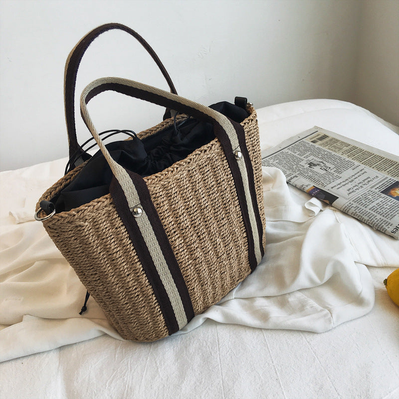 Woven shoulder straw bag with black and white strap on a bed with a newspaper.