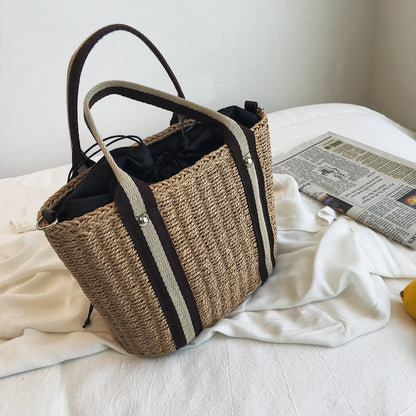 Woven shoulder straw bag with black and white strap on a bed with a newspaper.