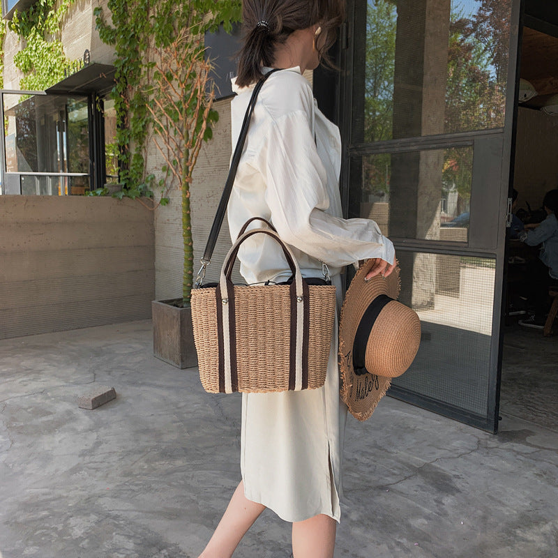 Woman walking with a woven straw shoulder bag with contrast handle and hat, standing in front of a glass door.