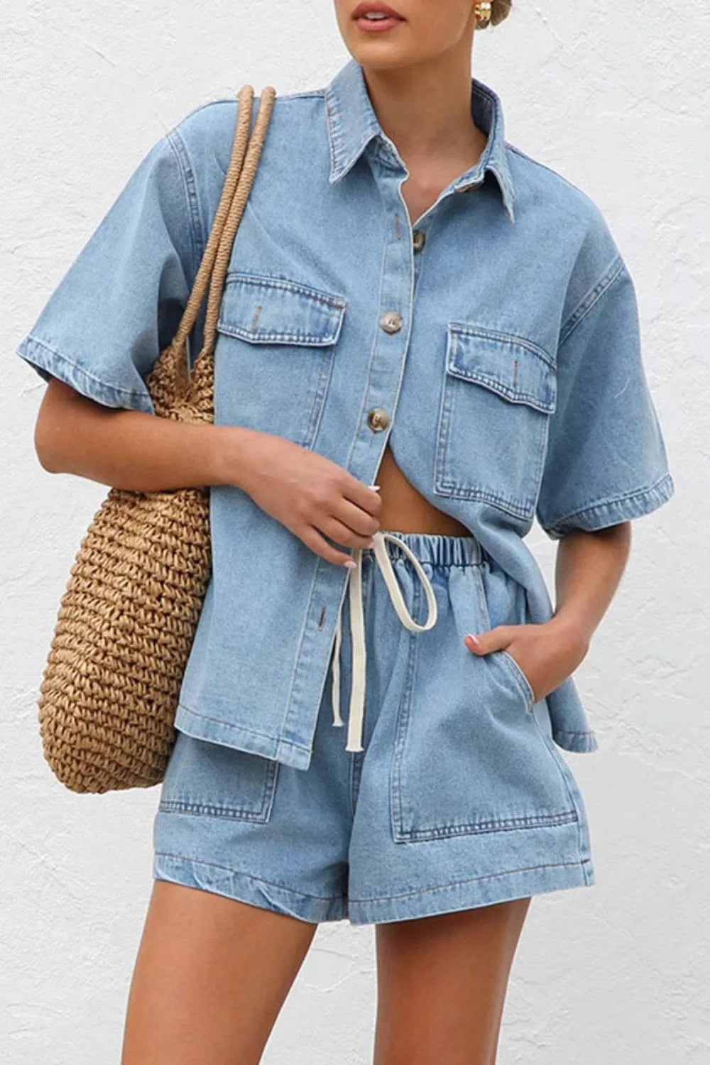 Woman wearing a light blue washed denim two piece shirt and shorts outfit, holding a woven bag against a white background.