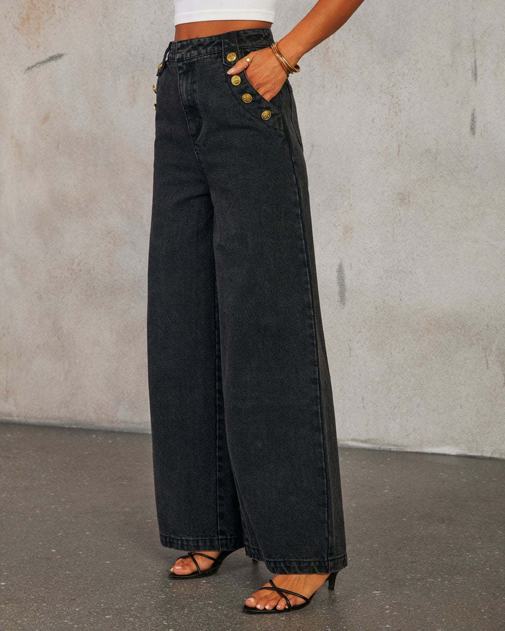 Side view of woman wearing high waist wide-leg black jeans with gold buttons against a plain background
