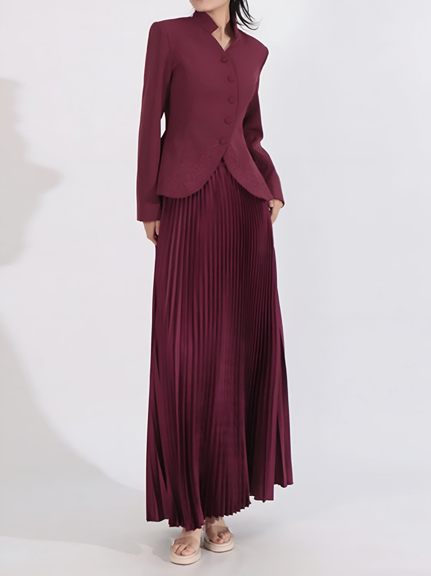 Woman posing in a structured embroidered jacket with pleated maxi skirt on a white background