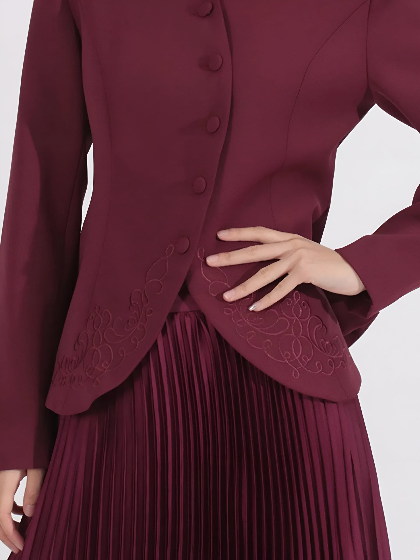  Woman wearing a  burgundy two piece co-ord with long sleeve embroidered detailed  jacket with pleated skirt on a white background