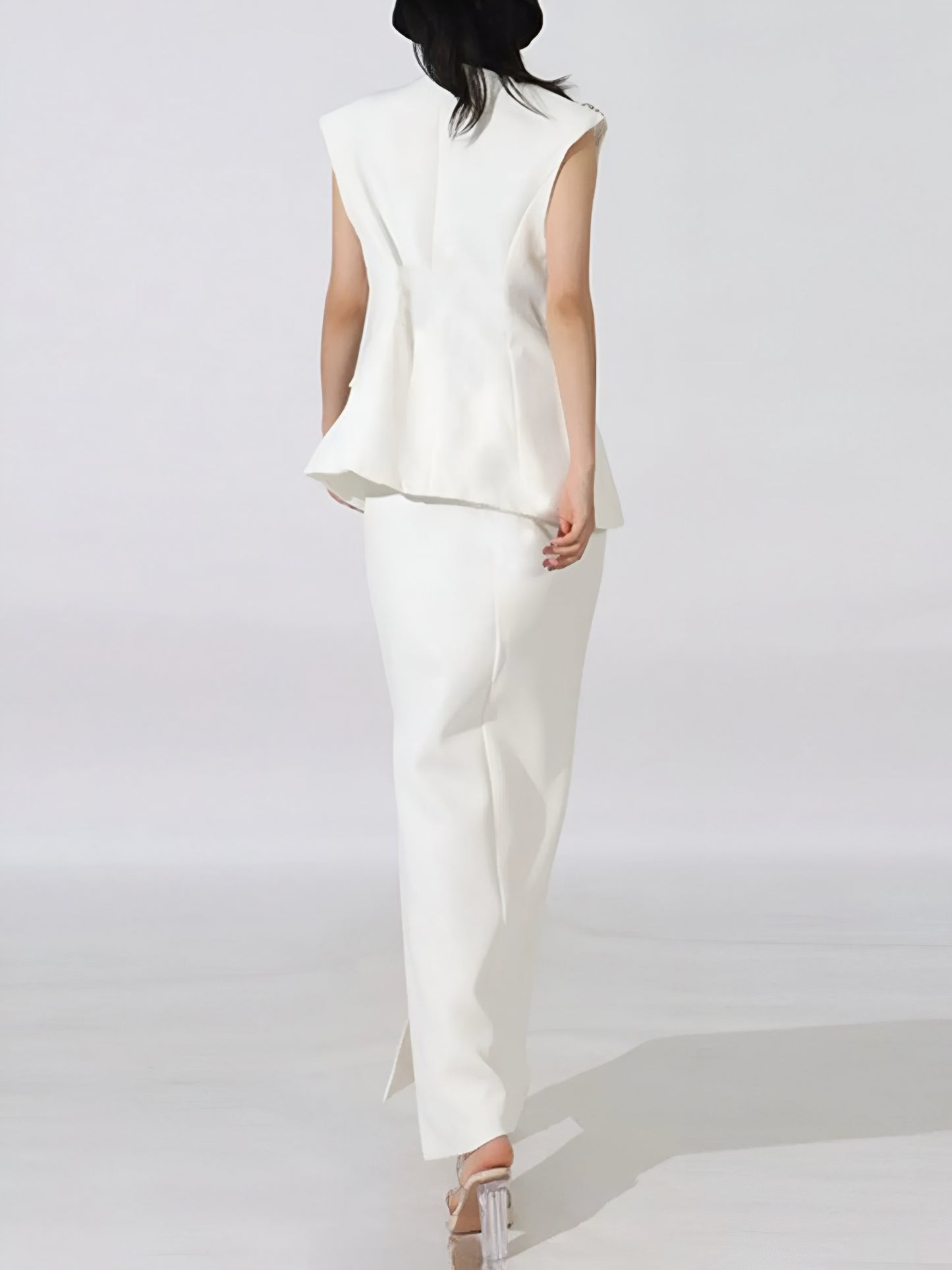 back view of woman wearing a white sleeveless high end luxury blazer top and maxi skirt with side slit on a plain background