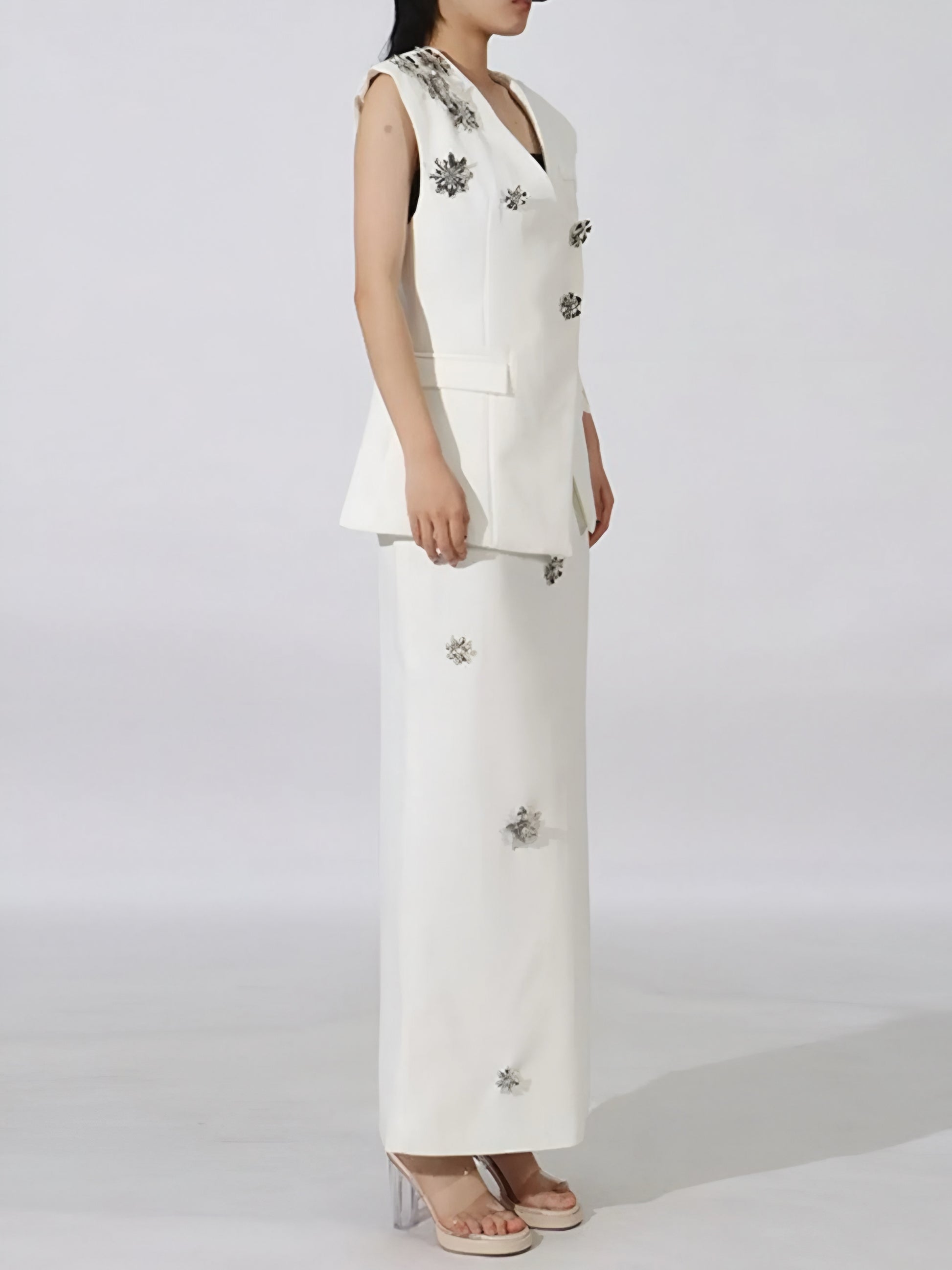 Side view of woman wearing a luxury floral sleeveless vest and maxi skirt set with floral embellishments on a plain background