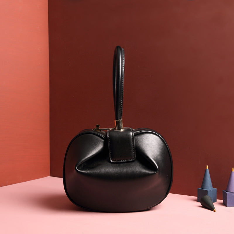 A black leather dumpling handbag with a buckle closure and a single handle, displayed against a two-tone background 