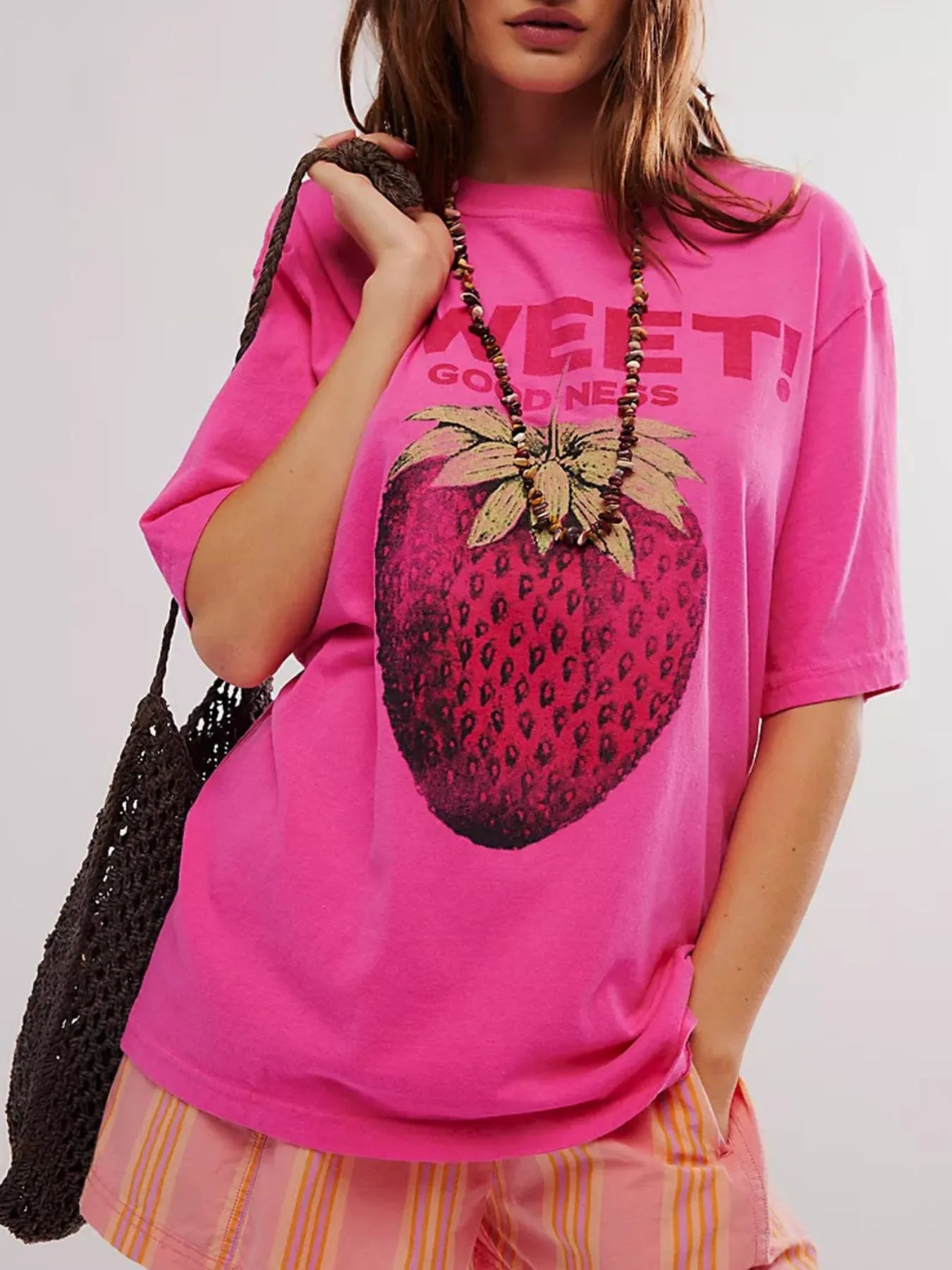 Front view of woman wearing a pink t-shirt with a strawberry graphic and text, holding a black bag.