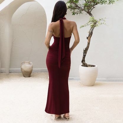 Back view of woman wearing a red/burgundy halter backless part maxi dress standing in an outdoor setting with a white wall and potted plant.
