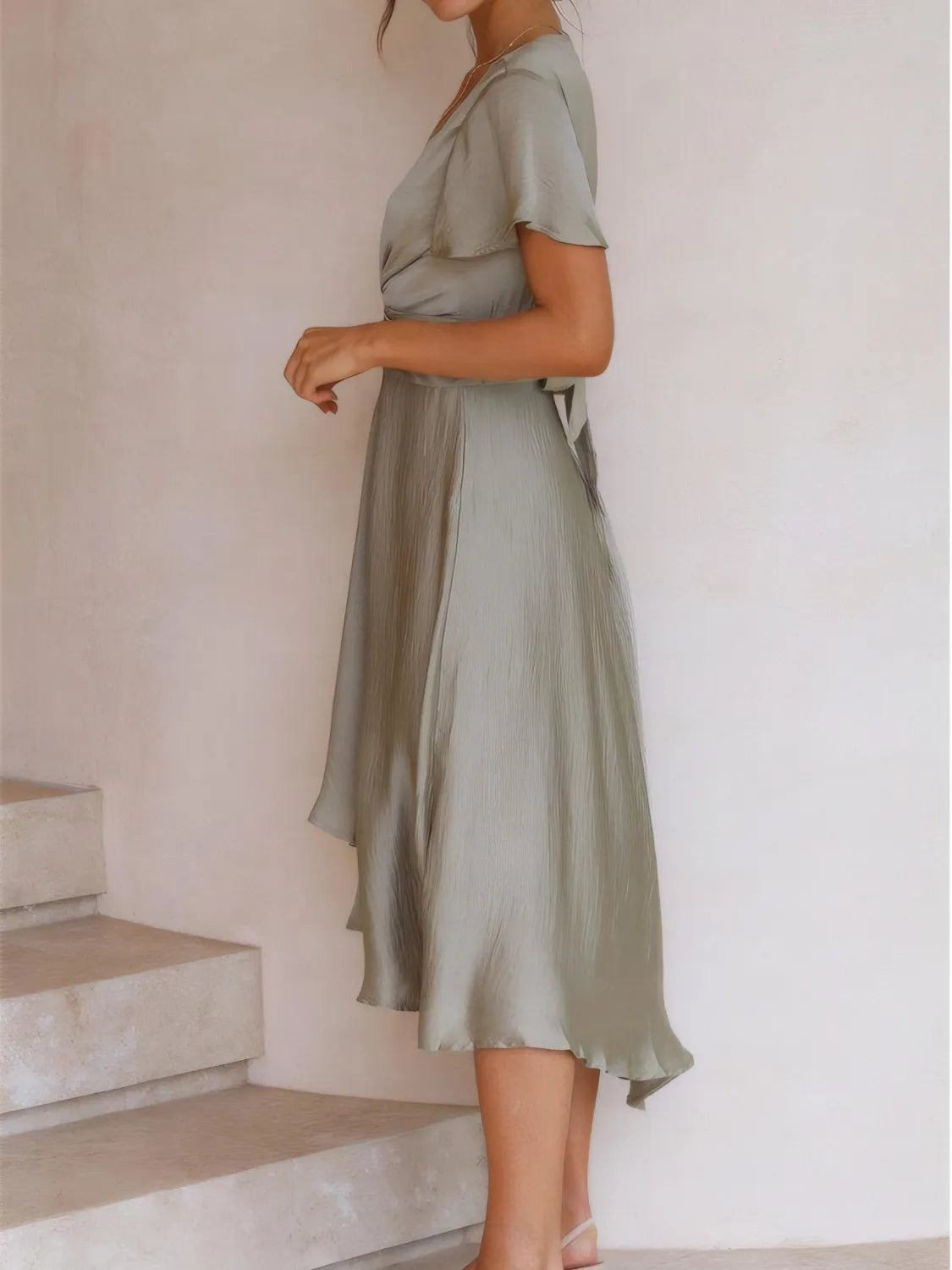 Model walking in sage high-low dress with flutter sleeves and deep v-neckline side view