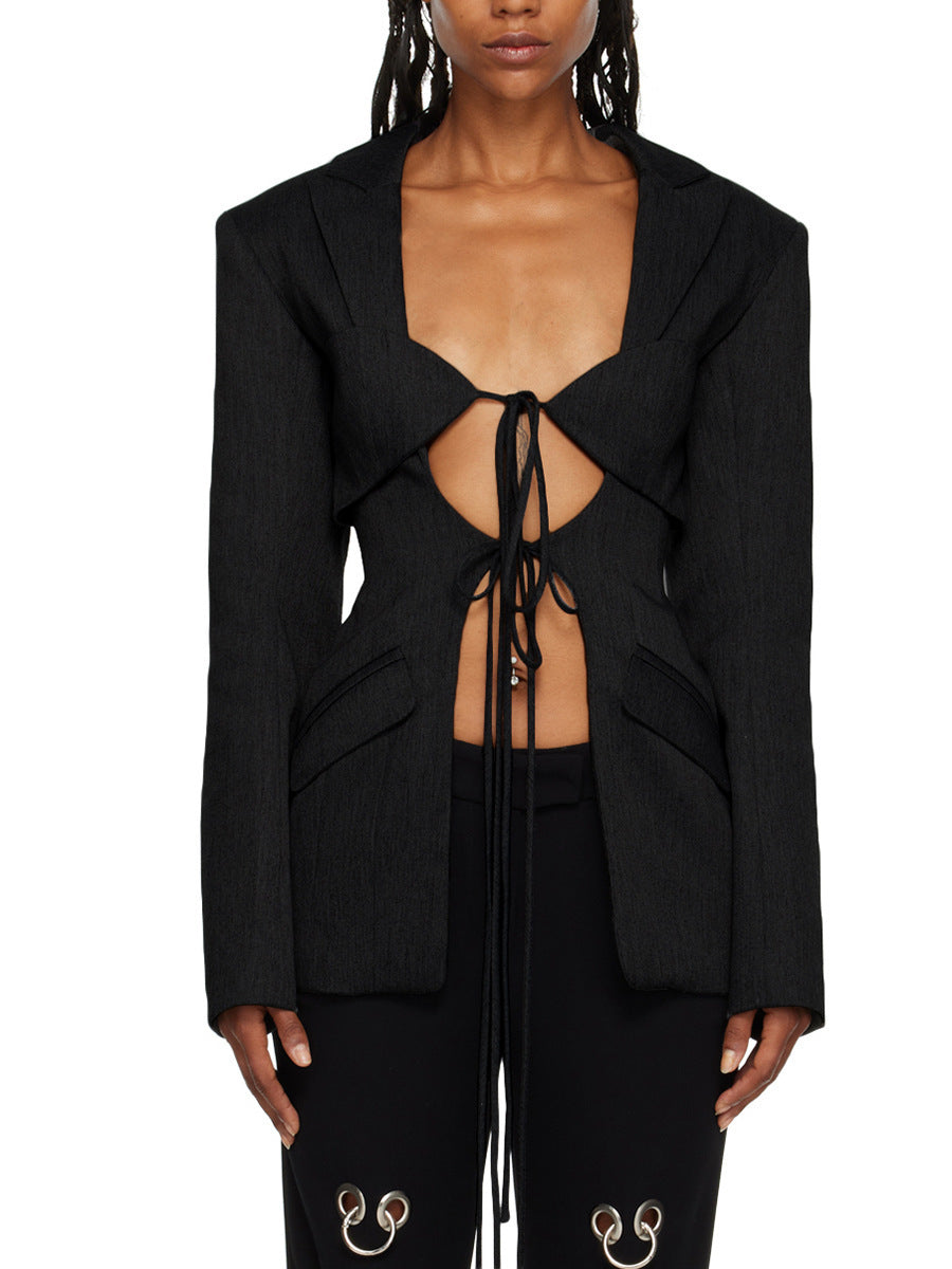 A woman wearing a black hollow-out cinched waist irregular long sleeve blazer with a deep v-neckline and lace up front detail paired with black pants featuring floral details.