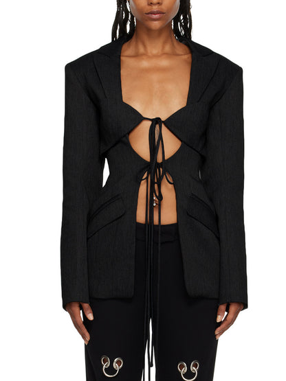 A woman wearing a black hollow-out cinched waist irregular long sleeve blazer with a deep v-neckline and lace up front detail paired with black pants featuring floral details.