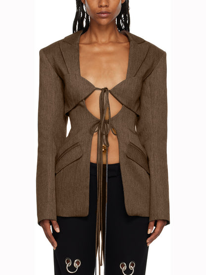A woman wearing a brown designer inspired hollow-out cinched irregular blazer with a deep v-neckline and long sleeves, paired with black pants