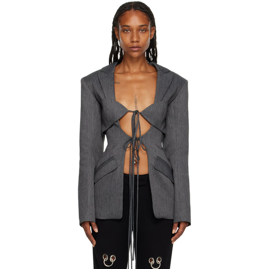A woman wearing a gray hollow-out cinched irregular blazer with a deep v-neckline and long sleeves, paired with black pants featuring floral details.