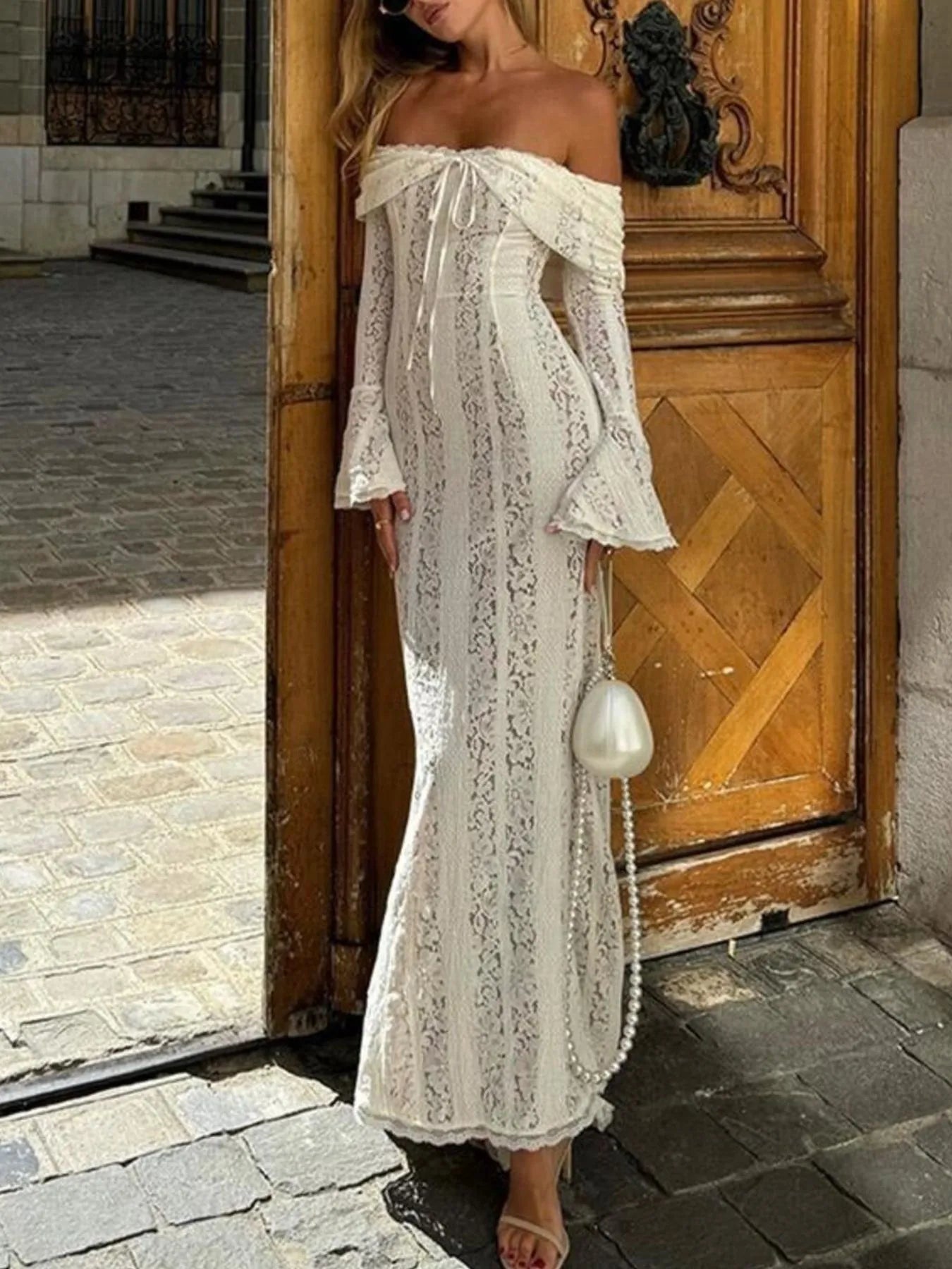 Woman in a white lace off shoulder maxi dress with flounce sleeves standing in front of a wooden door.