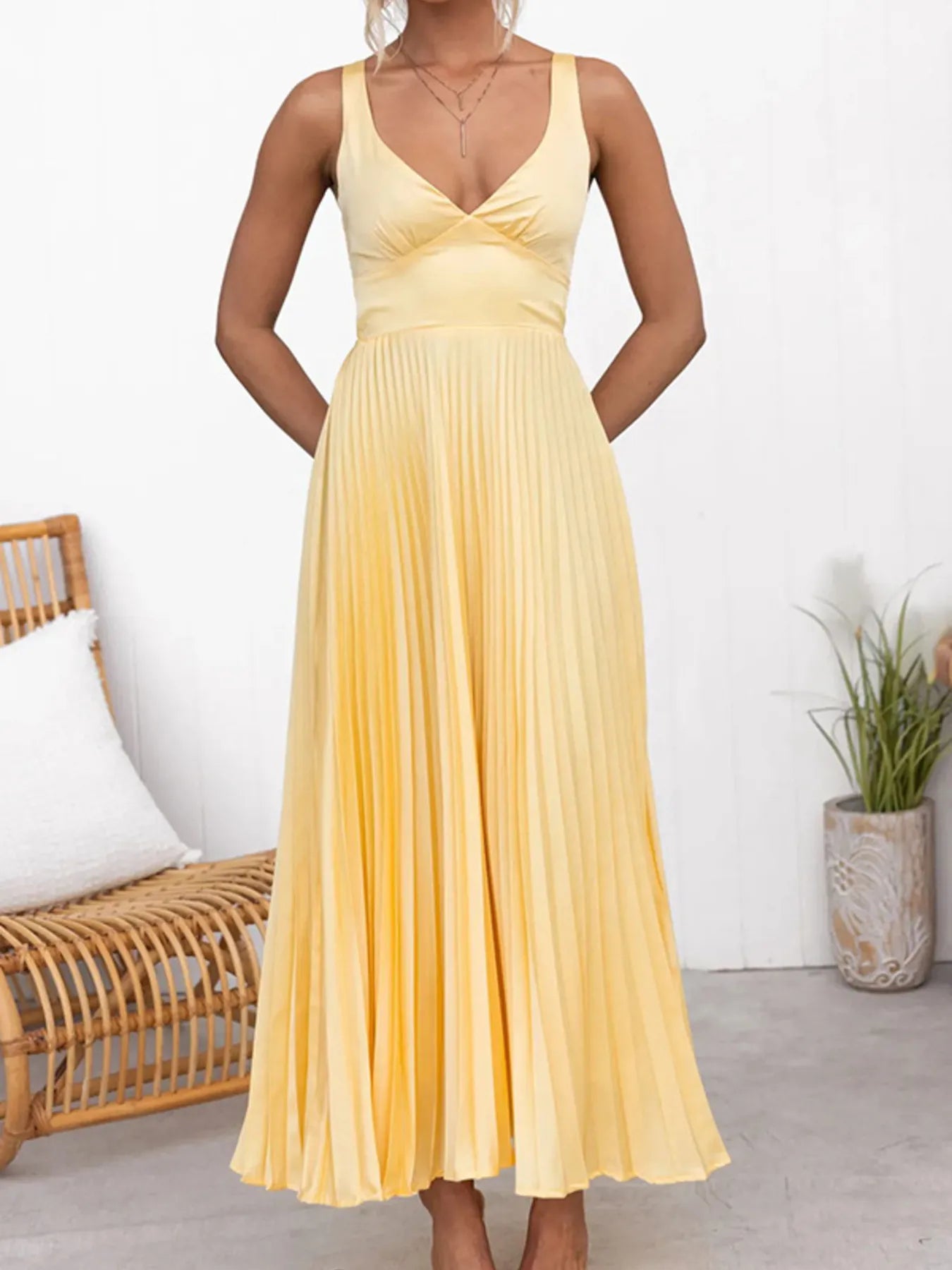 Woman wearing a yellow Sleeveless pleated maxi dress with V‑neckline in a room with a wicker chair and plant.