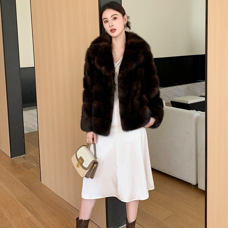 Woman wearing a high-fashion mid-length  brown faux fur coat in sable tone holding a purse in a room