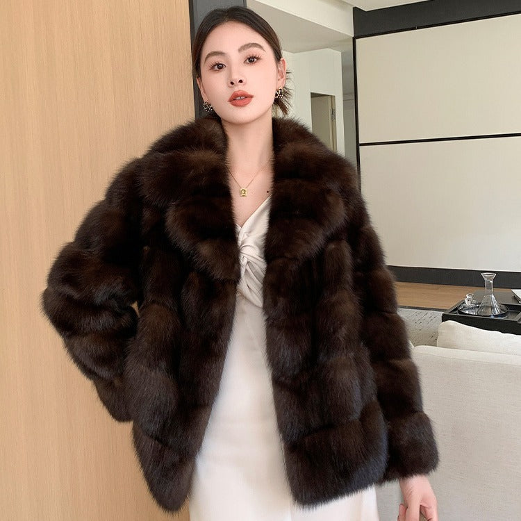 A woman wearing a brown artificial sable fur jacket with long sleeves, standing in a room with a wooden door