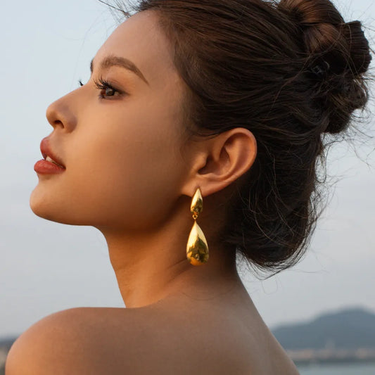 Woman wearing gold sculptural dangle drop earrings on a seaside background”

