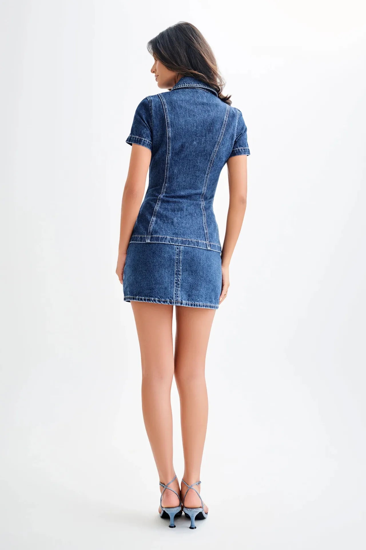 back view of woman wearing a two piece dark wash denim short sleeve top and mini skirt casual set on a white background