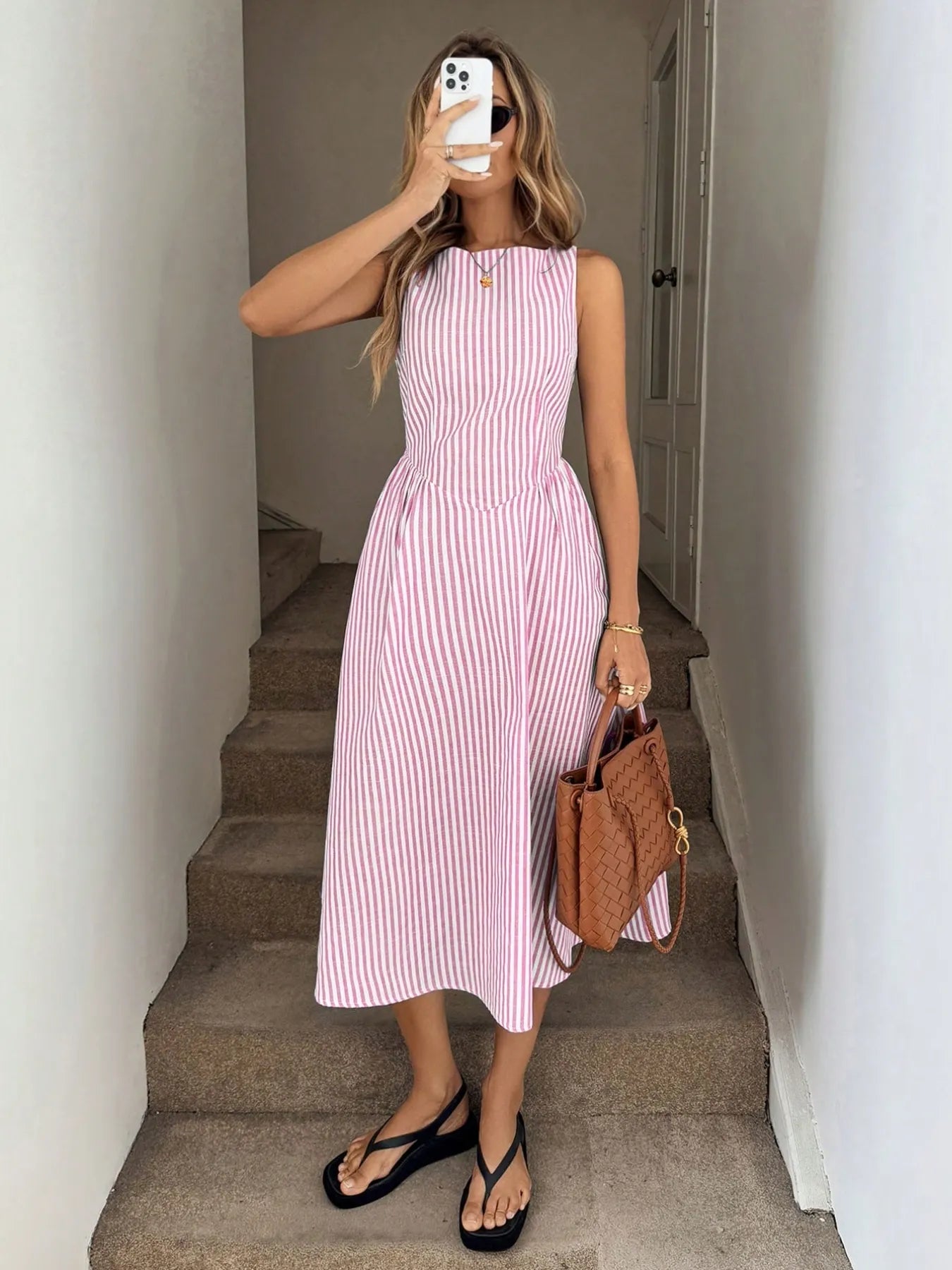 Woman wearing sleeveless pink striped midi dress outdoors holding a handbag and phone 