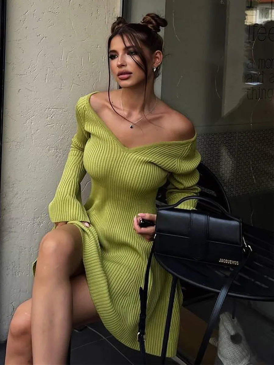 Woman in a green ribbed knit dress holding a black handbag against a neutral wall.