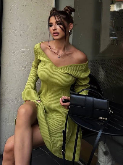Woman in a green ribbed knit dress holding a black handbag against a neutral wall.