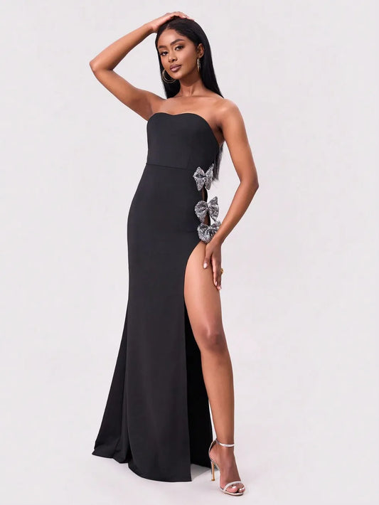 Model wearing a black strapless  silver bow trim maxi dress with high slit on a white studio background front view
