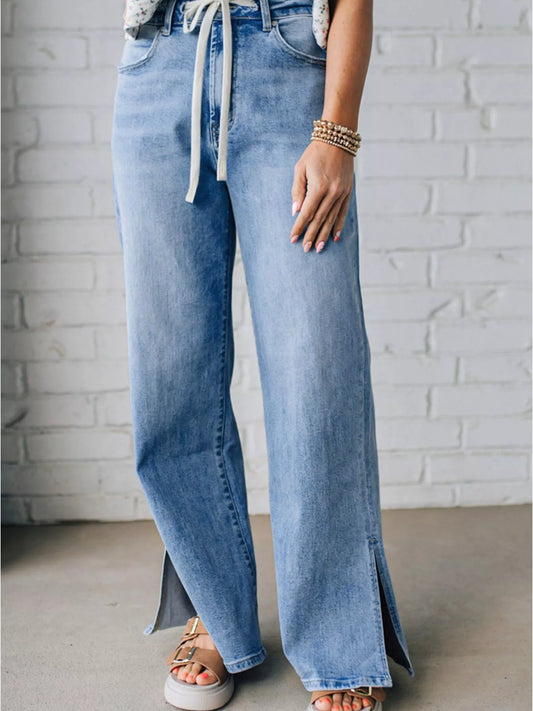 Woman wearing light blue high waist drawstring jeans with slit bottoms on a white brick wall background
