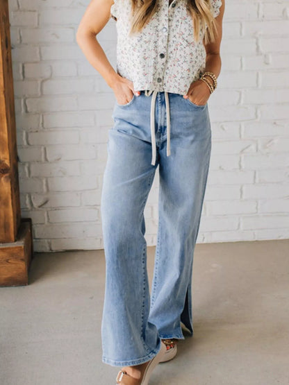 Woman wearing a floral top and light blue wash denim high waisted wide leg jeans with slits against a white brick wall.
