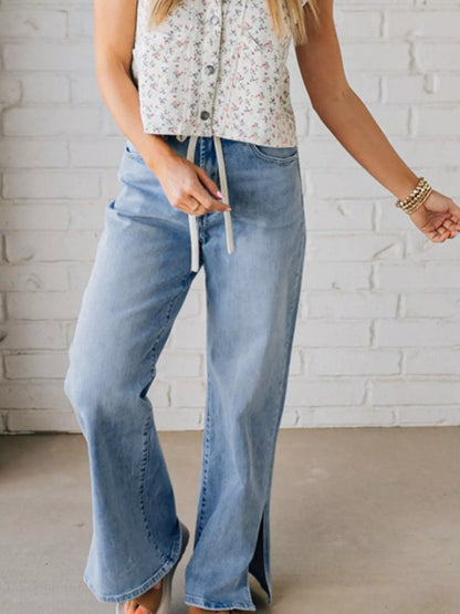 Woman wearing light blue drawstring denim high rise wide leg jeans and a floral top against a white brick wall.