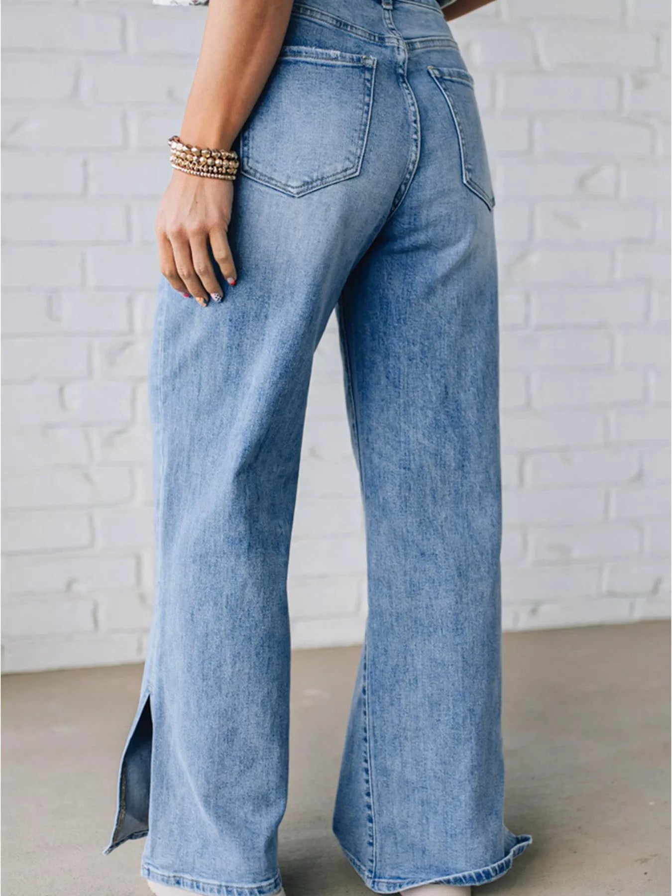 Back view of light blue high rise denim wide leg jeans with slit jeans worn by a woman against a white brick wall.