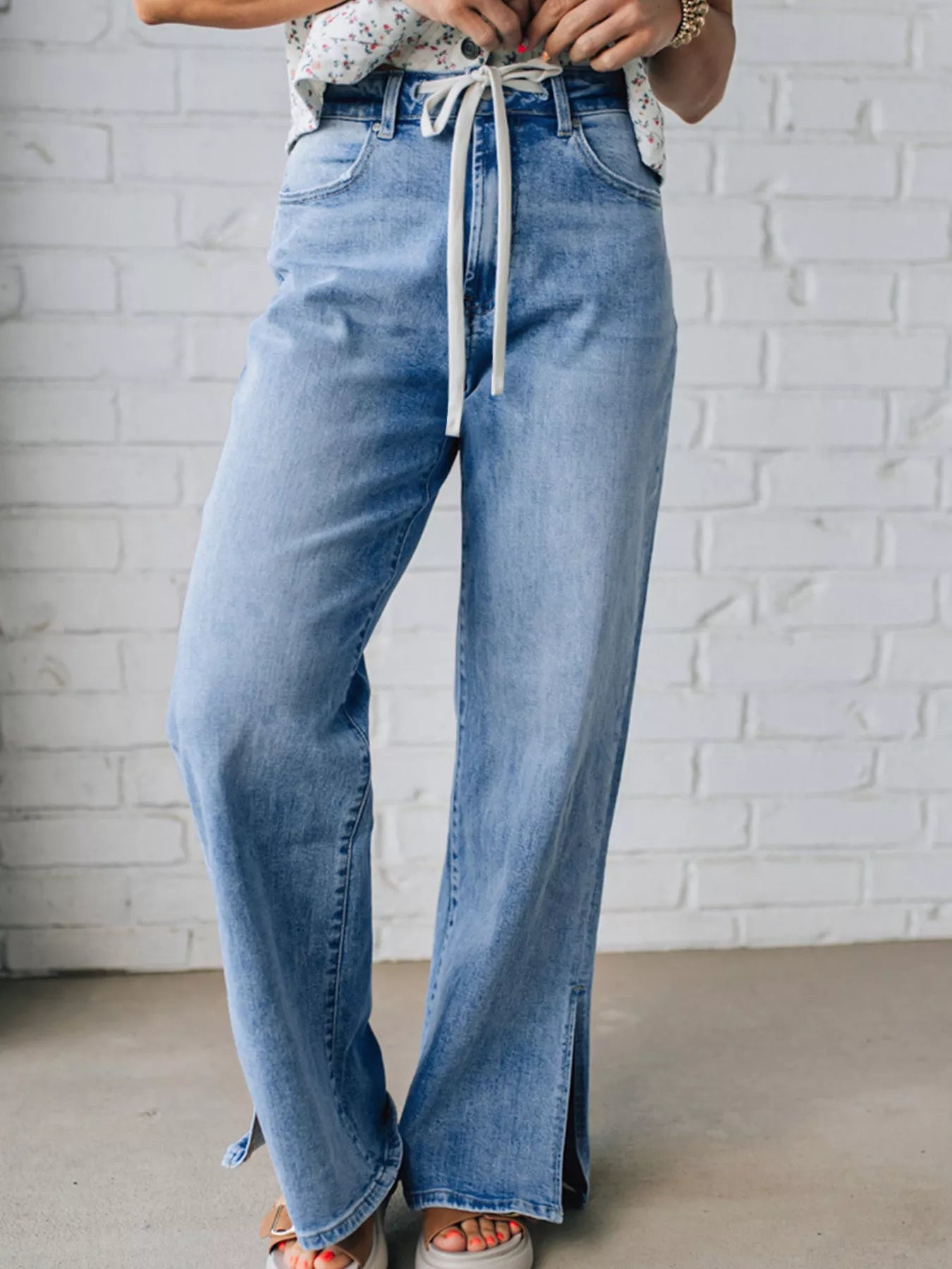 Light blue  drawstring jeans with slit legs worn by a woman against a white brick wall.