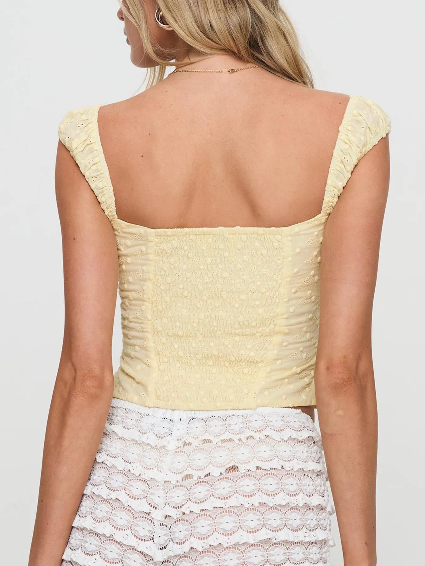 Back view of woman wearing a yellow Smocked Square Neck Top with cap sleeves and a white lace shorts on a plain background