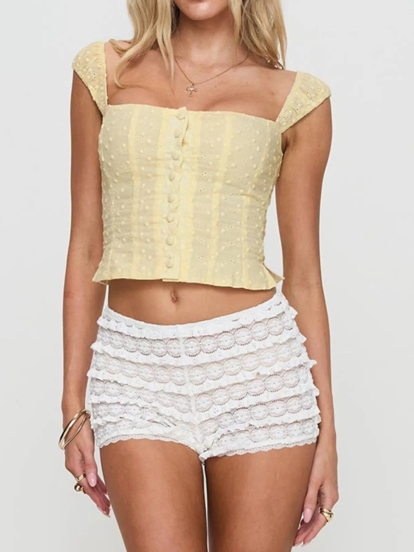 Woman wearing a yellow Smocked Square Neck Top with buttons and white lace shorts on a plain background