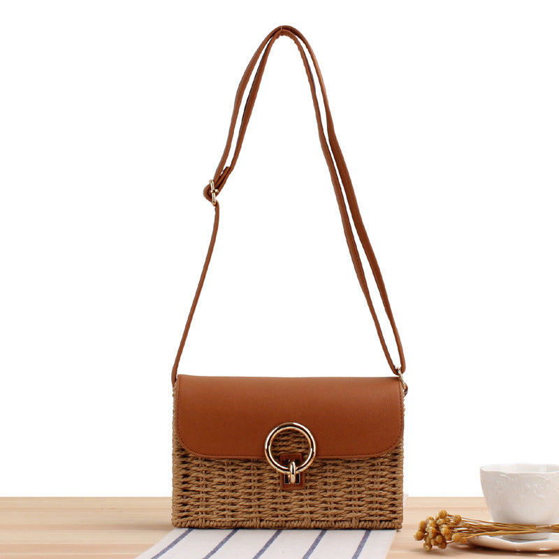Brown woven straw handbag with leather strap and flap on a light wooden surface.