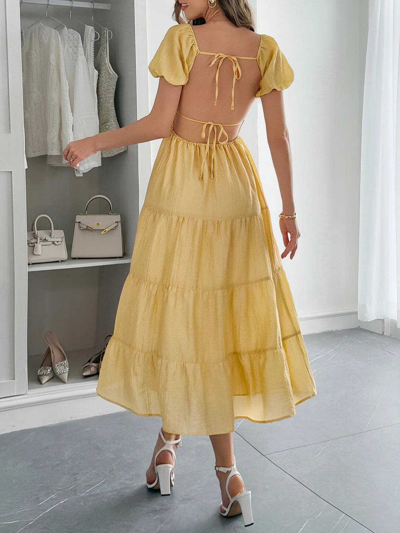 Back view of woman wearing a yellow Midi Dress with Short Sleeves  and tie up open back in a room with white walls and gray floor.