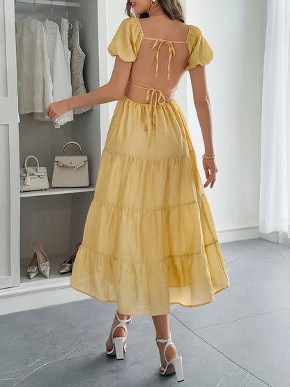 Back view of woman wearing a yellow Midi Dress with Short Sleeves  and tie up open back in a room with white walls and gray floor.